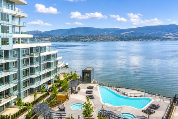 Modern living room with floor-to-ceiling windows overlooking coastal mountains and water, furnished with blue and cream sofas, wicker chairs, and potted plants.