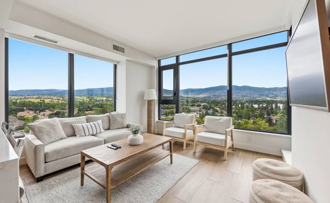 Modern living room with curved beige sofa, floor-to-ceiling windows overlooking lake and mountains, white walls, and light wood flooring.